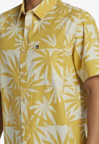 Camicia a maniche corte con bottoni di colore giallo, ornata da un grande motivo di foglie di palma bianche, dotata di taschino e leggeri bottoni in legno.