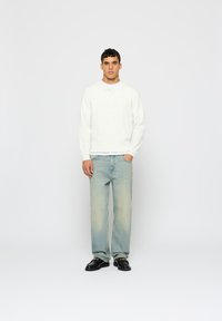 Fehér, hosszú ujjú henley póló gomb részletekkel, világoskék egyenes szárajú farmernadrággal és fekete mokaszinokkal párosítva. Egyszerű dizájn, laza szabás.