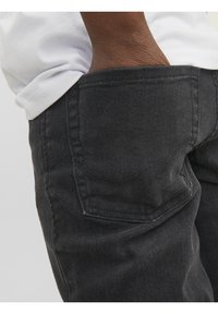 Main dans la poche arrière d'un jean en denim noir porté avec une chemise blanche, montrant la texture du tissu et les détails de la couture.