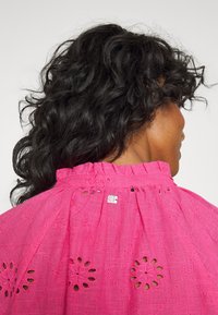 Blusa rosa bordada com decote franzido, apresentando padrões de ilhós e uma pequena etiqueta de logotipo na parte de trás. Tecido leve e texturizado.