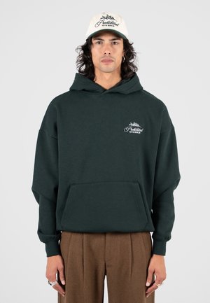 Hombre con cabello hasta los hombros que lleva una sudadera verde oscura con bolsillo delantero y pantalones marrones, complementado con anillos y una gorra blanca.