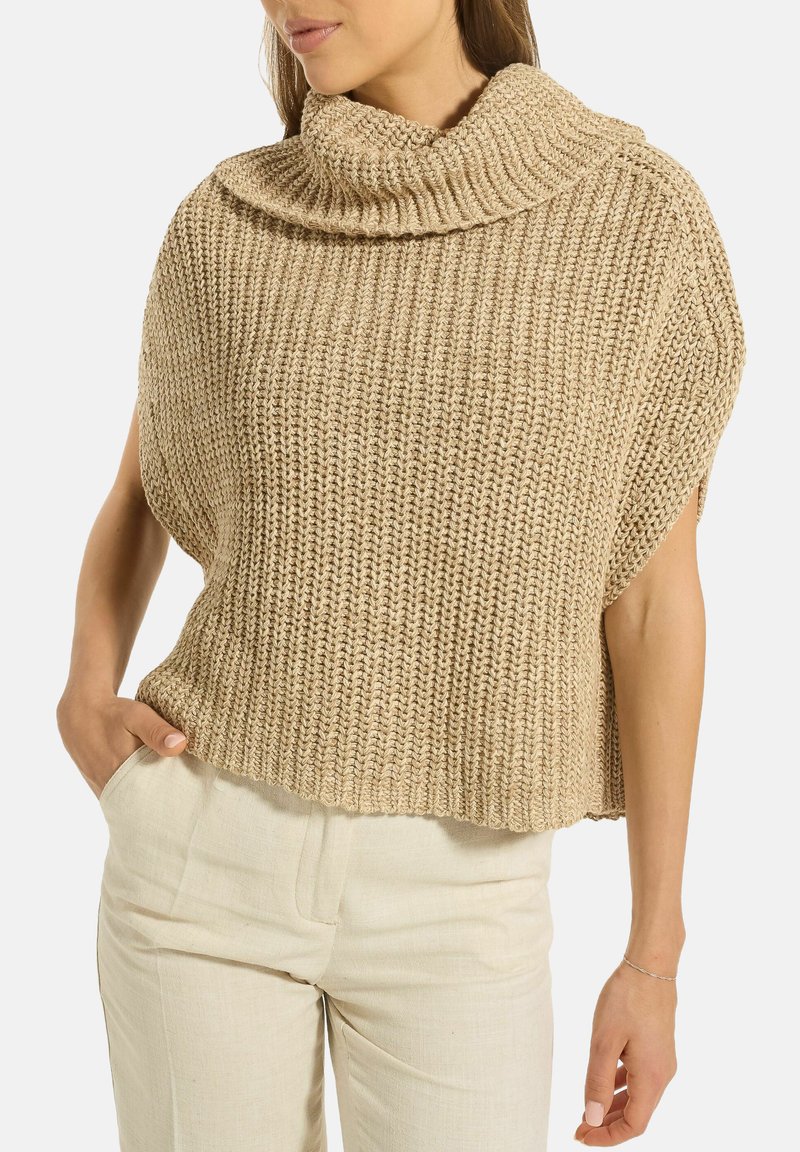 Frau trägt einen beigen, grob gestrickten ärmellosen Pullover mit einem umgeschlagenen Rollkragen, kombiniert mit hellen, hochgeschnittenen Hosen.