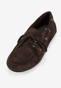 Zapato mocasín de ante marrón con punta redonda, detalle de cordones y ojales de metal, que presenta una textura suave y suela de goma plana.