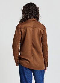 Giacca marrone in suede con una vestibilità su misura, dotata di cucitura sulla parte posteriore e polsini abbottonati. Indossata sopra pantaloni blu, vista da dietro.