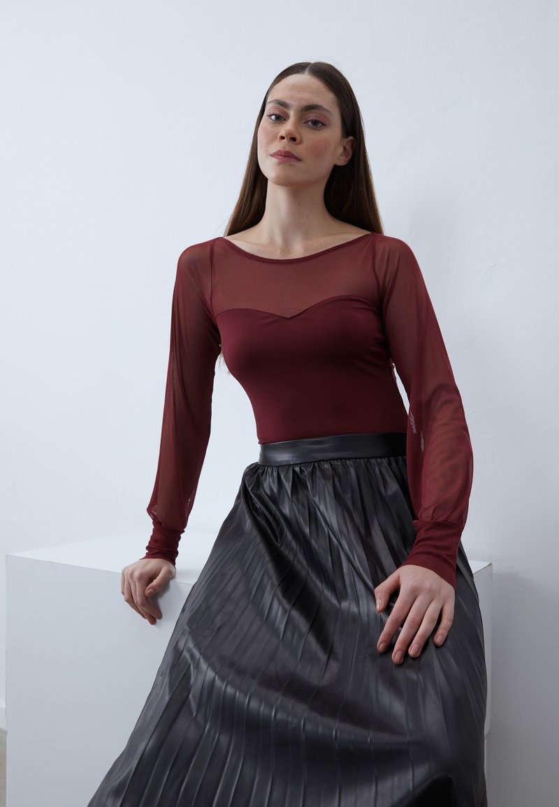 Burgundy långärmad topp med genomskinlig design och hjärtformad halsringning, kombinerad med en veckad svart kjol med en slät textur.