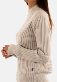 Maglione beige a coste con colletto alto, caratterizzato da un design a maglia cable e un piccolo logo metallico sul lato. Maniche lunghe. Trama morbida.