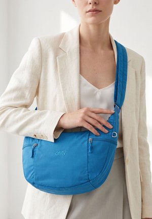Vrouw met een crèmekleurige blazer en wit topje houdt een blauwe crossbodytas met ritszakken en "GMV" reliëf aan de voorkant.