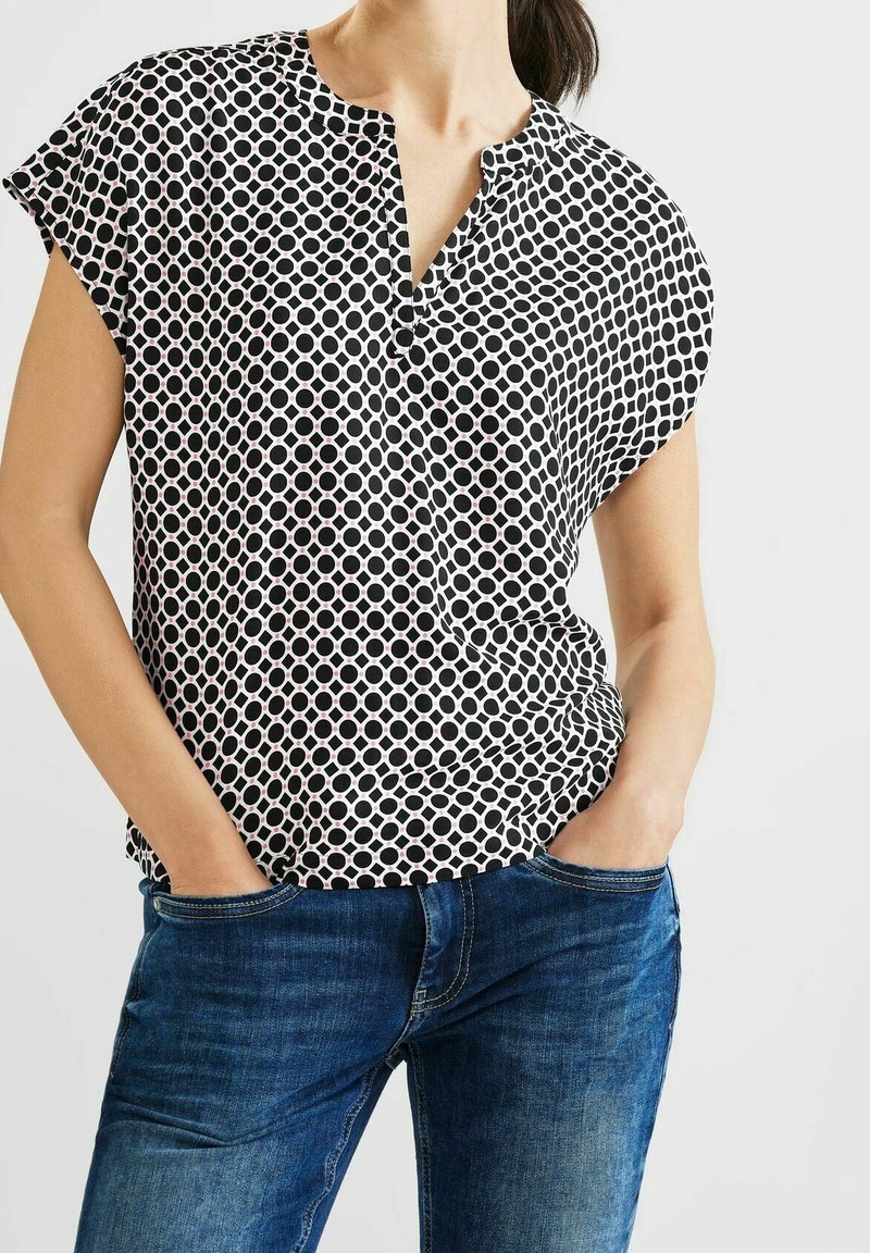 Czarna i biała bluzka z krótkim rękawem i okrągłym dekoltem, ozdobiona wzorem w okręgi. Noszona z niebieskimi dżinsami, podkreślającymi swobodny krój.