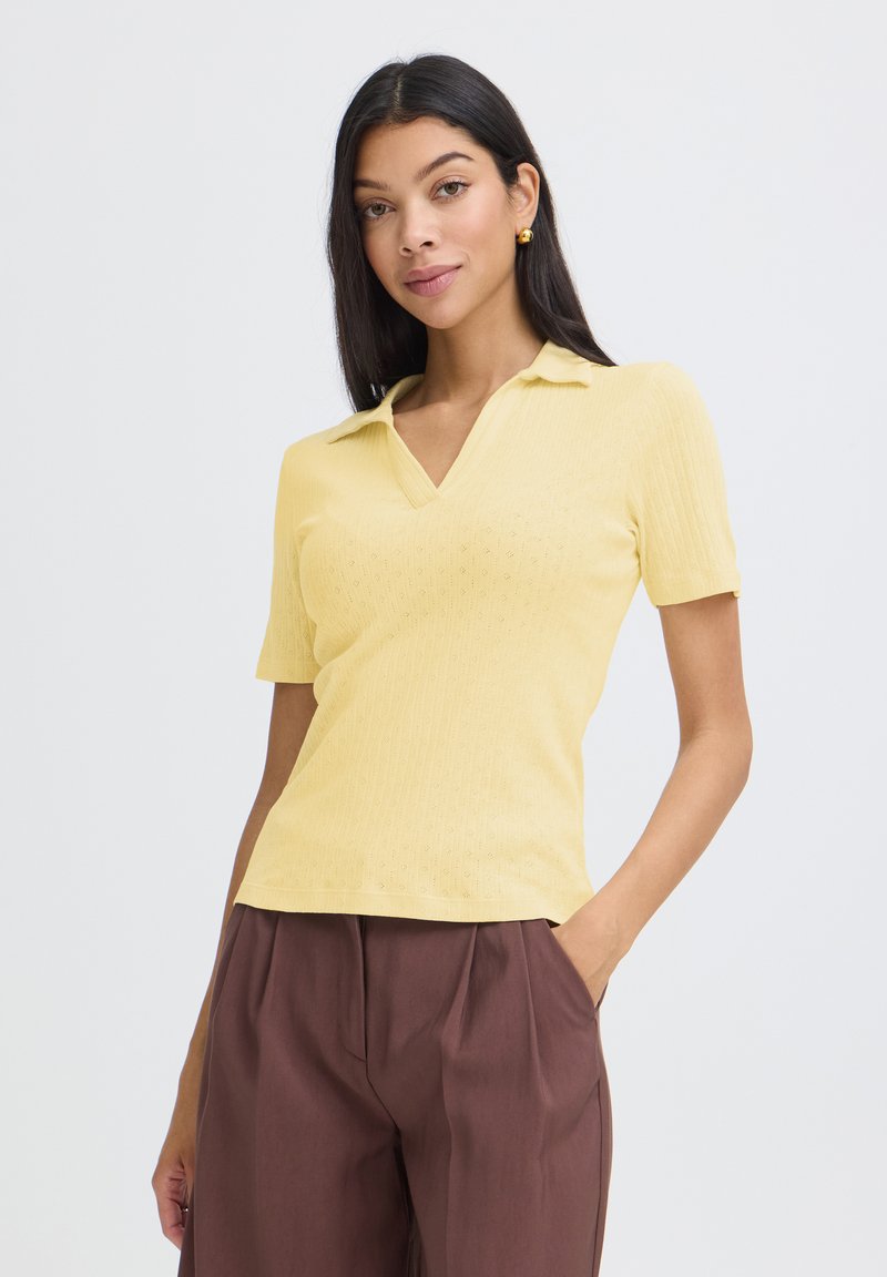 Femme aux longs cheveux foncés portant un polo jaune à manches courtes et un pantalon marron taille haute, debout avec une main dans la poche.