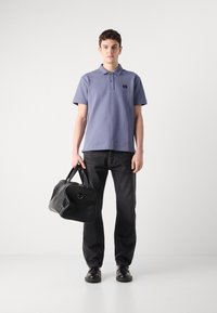 Polo shirt violet clair avec un col et un logo, associé à un jean noir. Le modèle tient un sac de sport en cuir noir, se tenant sur un fond neutre.