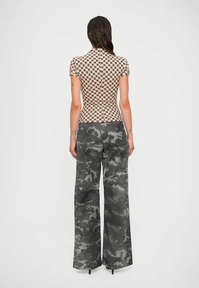Frau mit langen, dunklen Haaren, die ein beigefarbenes, gemustertes Kurzarmtop und graue Camouflage-Hosen mit weitem Bein trägt, stehend vor einem schlichten weißen Hintergrund.