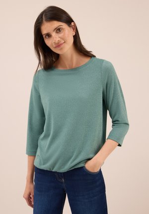 Femme aux longs cheveux bruns, portant un haut vert étincelant à manches trois quarts et un jean bleu foncé, se tenant avec une main dans la poche.