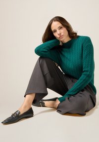 Türkisfarbener Kabelstrickpullover mit Rundhalsausschnitt, kombiniert mit grauen, weit geschnittenen Hosen und schwarzen flachen Schuhen mit goldenen Akzenten.