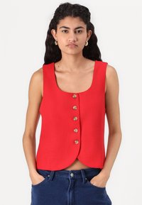 Red sleeveless top with a rounded neckline and five button closure down the front, made from a textured fabric, paired with dark blue jeans.