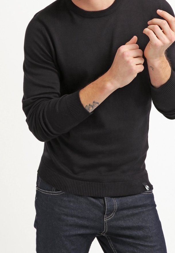 Schwarzer Strickpullover mit Rundhalsausschnitt und gerippten Bündchen, kombiniert mit dunklen Jeans. Hat ein Logo am unteren Saum.