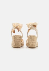 Sandales compensées beiges avec une texture en paille tressée et un doux nœud en tissu à l'arrière. Semelle lisse avec une hauteur modeste.