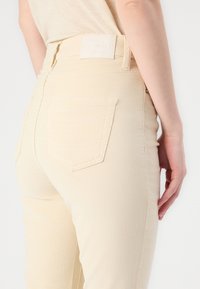 Jean beige à coupe ajustée, avec des poches arrière, une texture lisse et une étiquette de marque sur la ceinture.