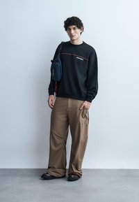 Jonge man staat tegen een effen muur, draagt een zwart sweatshirt, losse bruine broek, zwarte schoenen en een marineblauwe crossbody tas.