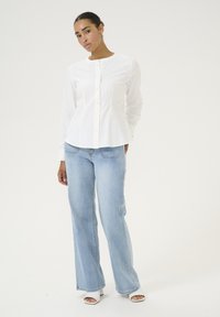 Chemise blanche à manches longues avec un col rond, portée avec un jean ample bleu clair et des talons ouverts blancs sur un fond neutre.