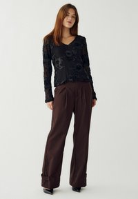 Zwarte lange mouwen top met bloemenborduursel, gecombineerd met bruine geplooide broeken en zwarte hoge hakken. Glad materiaal, relaxte pasvorm.