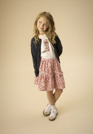 T-shirt blanc avec un motif de botte de cow-boy, superposé avec un cardigan foncé. Jupe à volants florale dans des tons roses. Baskets blanches avec des accents tan.