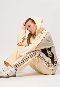 Fiatal nő bézs kapucnis melegítőben, fekete és piros logós csíkokkal, a padlón ül, fejét a kezére támasztja, elgondolkodva néz.