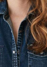 Närbild av en person med långt rött hår iförd en delvis knäppt blå denimjacka.