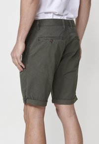 Vīrietis, tērpies tumši zaļos, iegriezumos shorts un baltā kreklā, redzams no aizmugures, ar roku atpūšoties pie sāna.