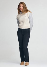 Cremfarbener Strick-Weste mit einem strukturierten Zopfmuster über einem blau gestreiften Hemd, kombiniert mit dunklen Hosen und hellbraunen Mokassins.