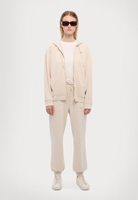 Bež hoodie s vrvicami in stranskimi žepi, v kombinaciji s prilegajočimi hlačami in belimi teniskami. Model nosi črne pravokotne sončne očala.