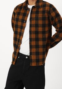 Hnědá a černá kostkovaná flanelová košile s knoflíky, s límcem a jednou náprsní kapsou, nošená přes bílé tričko a černé kalhoty.