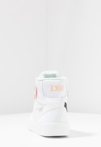 Korkea valkoinen lenkkari, jossa kangasosa, oranssi logo kantapäällä, kontrastoiva musta yksityiskohta sivulla ja teksturoitu välikansi.