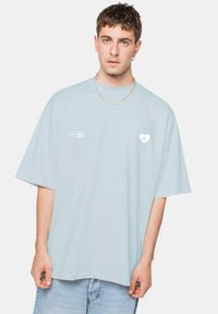 Camiseta de gran tamaño de color azul claro, hecha de algodón. Presenta mangas cortas, cuello redondo y un pequeño logo de corazón blanco con "EK" en el lado izquierdo del pecho.