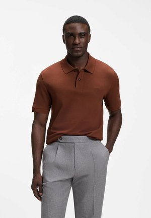 Man die een bruin poloshirt met korte mouwen en lichtgrijze, hoge taillebroek draagt, staand tegen een effen witte achtergrond.