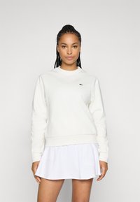 Kremfarget sweater med rund hals og ribbestrikkede mansjetter, med en liten grønn logo på brystet, kombinert med et hvitt plisséskjørt.