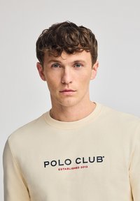 Sudadera crema con cuello redondo acanalado, que presenta el texto "POLO CLUB" en azul marino y rojo. Textura suave y corte regular. Fondo liso.