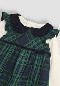 Vestido de cuadros verdes y marinos con una capa inferior blanca de mangas largas, que presenta un cuello tipo Peter Pan de terciopelo marino y mangas con volantes.