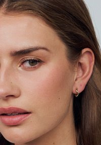 Guldsmycken i vågform med en slät textur, bärs i en models öra, kompletterat med naturlig makeup och rakt hår.