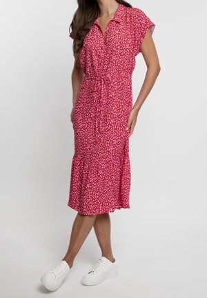 Kobieta ubrana w czerwoną sukienkę midi z różowymi, abstrakcyjnymi plamami, krótkimi rękawami, przewiązaną w talii, w białych sneakersach na platformie na jednolitym tle.