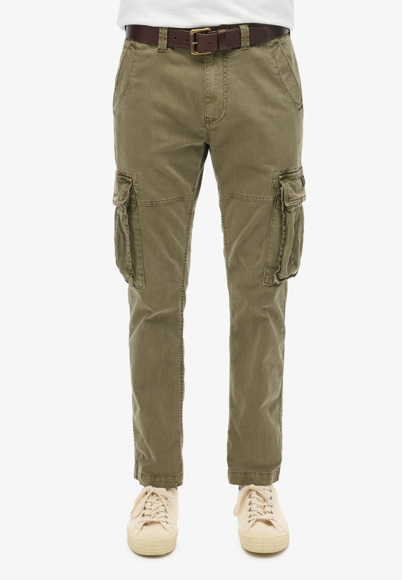 Pantalones cargo verde oliva con bolsillos laterales, cremallera y presillas para cinturón, fabricados en suave tejido de algodón, combinados con zapatillas beige claras.
