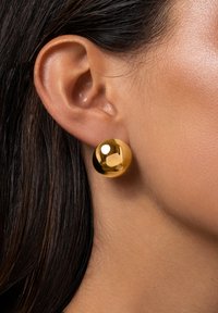 Orecchini a perno in metallo color oro con un design rotondo e liscio, che riflette la luce, indossati vicino al lobo dell'orecchio. Aspetto minimalista e lucido.