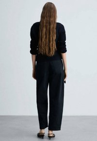 Schwarze, weit geschnittene Jeans werden mit einem marineblauen Strickpullover kombiniert. Langes, welliges, hellbraunes Haar fällt den R�ücken hinunter. Schwarze Sandalen vervollständigen das Outfit.