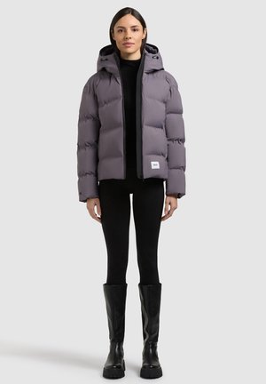 Mujer con chaqueta acolchada con capucha gris, leggings negros y botas negras hasta la rodilla, de pie frente a un fondo liso y claro.