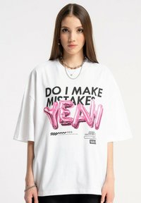 Młoda kobieta z długimi brązowymi włosami nosi białą, oversized koszulkę z napisem "CZY POPEŁNIAM BŁĘDY, TAK" w różowych, balonowych literach.