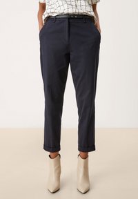Pantalons habillés navy avec une coupe ajustée, ourlets retroussés et passants de ceinture ; associés à des bottines beiges à fermeture éclair sur le côté.