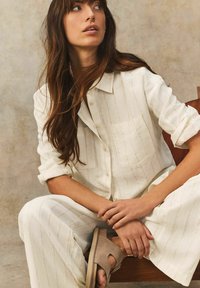 Witte linnen blouse met lichte verticale strepen en een borstzak, gecombineerd met bijpassende lichte broek en taupe open teen sandalen.