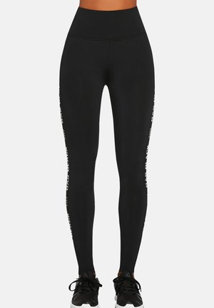 Kobieta nosząca czarne legginsy z wysokim stanem z białym napisem po bokach oraz czarne buty sportowe, stojąca na jednolitym tle.