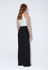 Woman with long red hair wearing a white sleeveless top and high-waisted wide-leg black pants, standing facing a white wall.