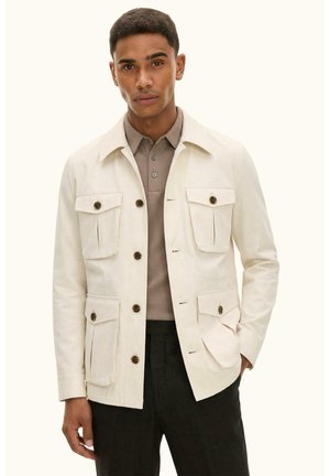 Mand iført beige jakke med knapper og fire forlommer over en taupe polo og sorte bukser, stående mod en ensfarvet baggrund.