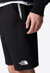 Pantalones cortos negros de tela suave, con una etiqueta de logo blanca, bolsillos laterales y una cinturilla con cordón y detalles de marca.